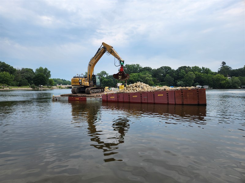 An excavator on a floating platform moves building materials in Fountain Lake.